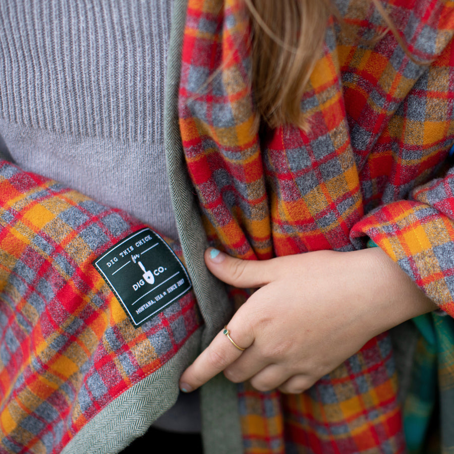 A close-up of a red plaid camp blanket that is wrapped around a persons shoulders. The Dig + Co. logo is displayed on the blanket. 