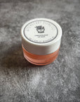 A closed glass pot of cardamom rose lip salve. The lid is white and displays the Apothecary Esthetics logo. 