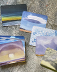 Watercolor illustrations on the back of the cards from the cloud spotter deck displayed on a gray surface. 