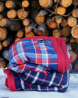 Blue and red plaid flannel Camp Blanket folded and placed in the snow in front of a wood pile.