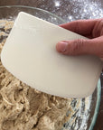 A hand holds a white bowl scraper over a glass bowl full of dough