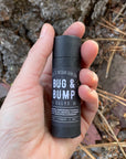 A hand holds a black tube of bug & bump salve in front of gray rocks covered in pine needles.