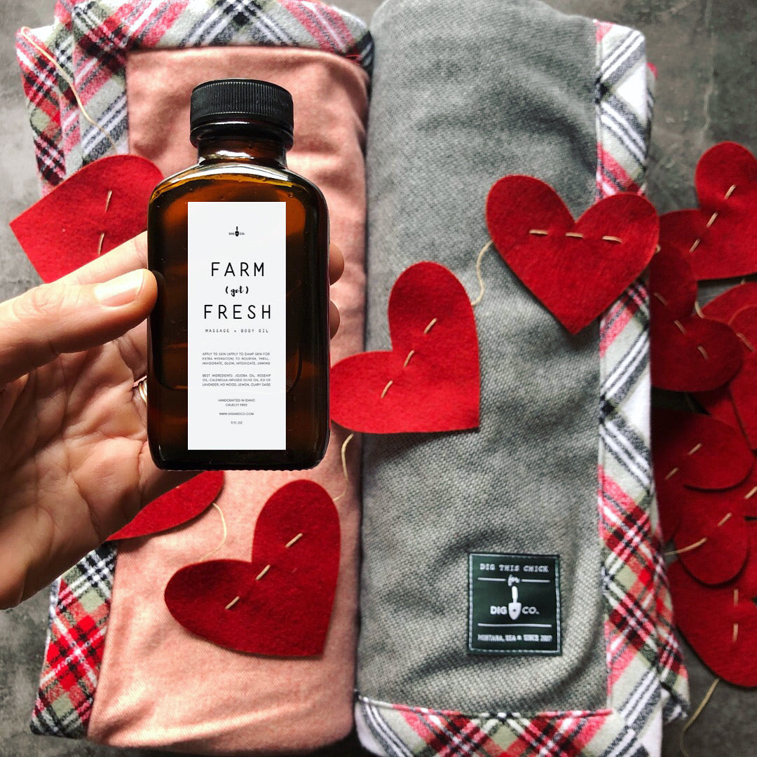 A hand holds a brown glass jar of massage oil in front of two rolled gray and pink blankets. Fabric hearts on a string are draped on top of the blankets. 