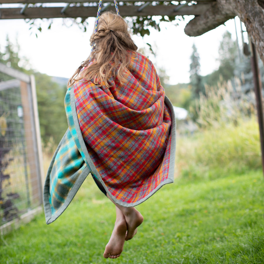 A young barefoot girl with loose blonde hair sits on a swing over green grass wrapped in a flannel blue and red camp blanket. 