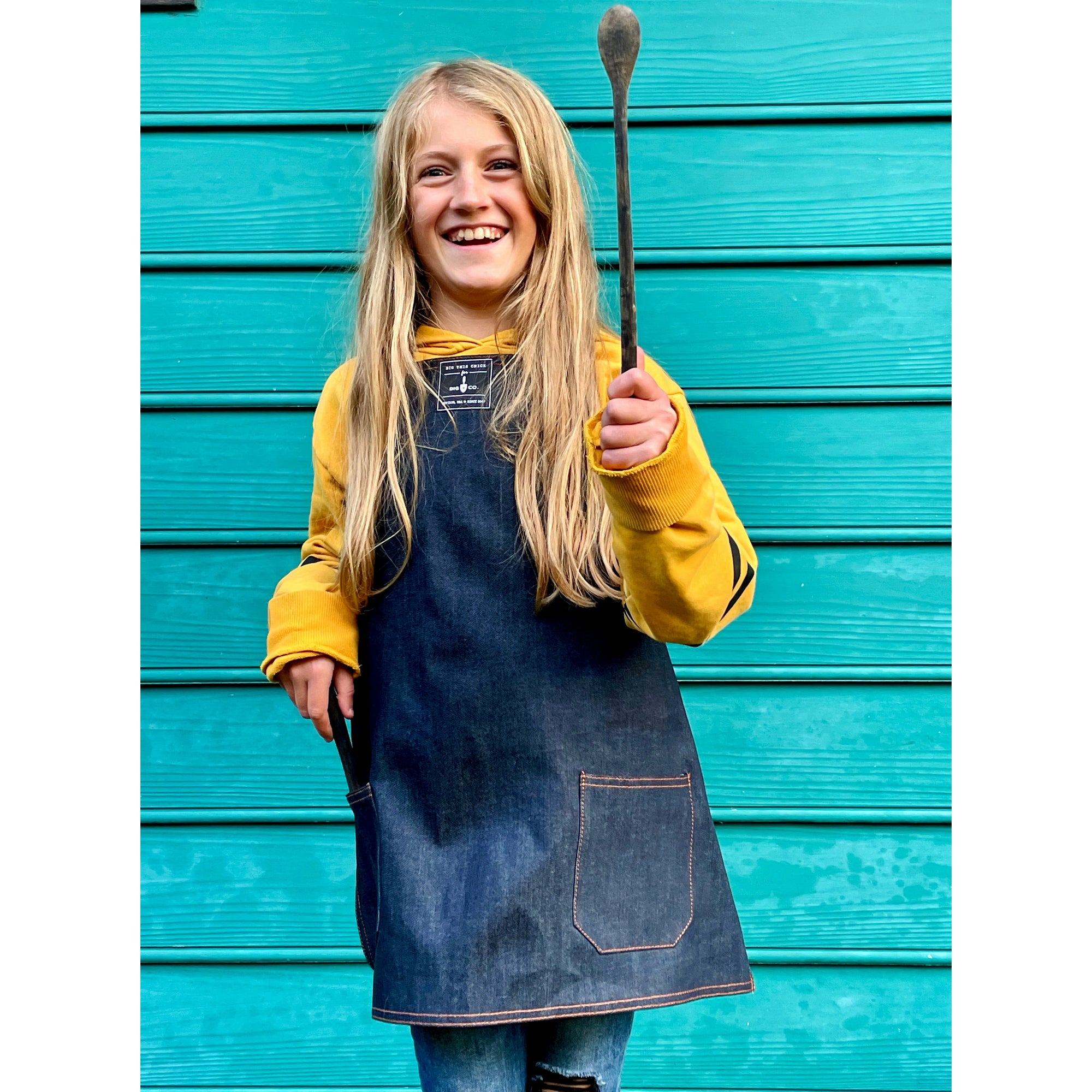 A child with long blonde hair wears the denim child's apron and holds up a wooden spoon. 