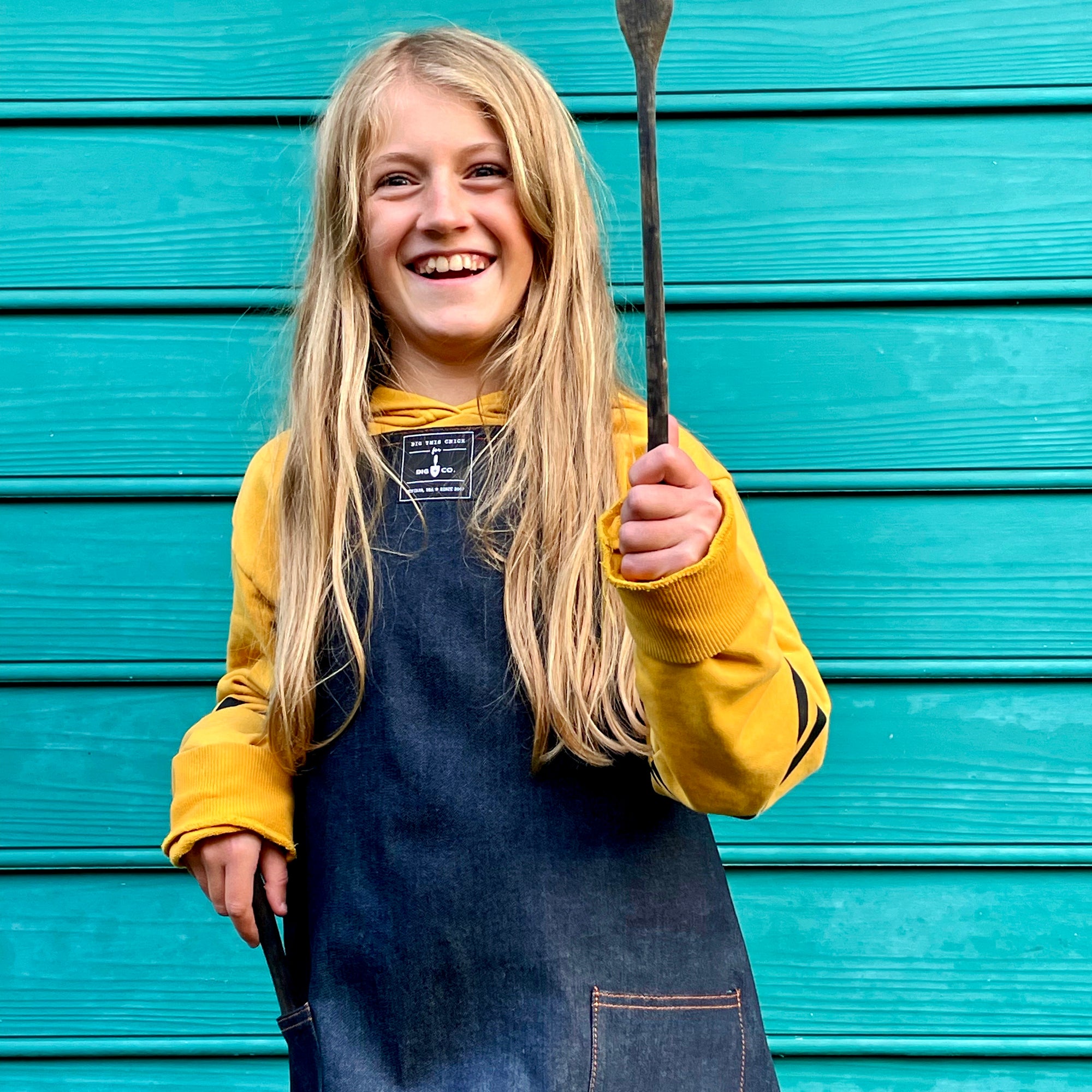 A child with long blonde hair wears the denim child&#39;s apron and holds up a wooden spoon. 