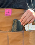 A person wears the canvas utility apron and pulls their phone out of the front pocket. 