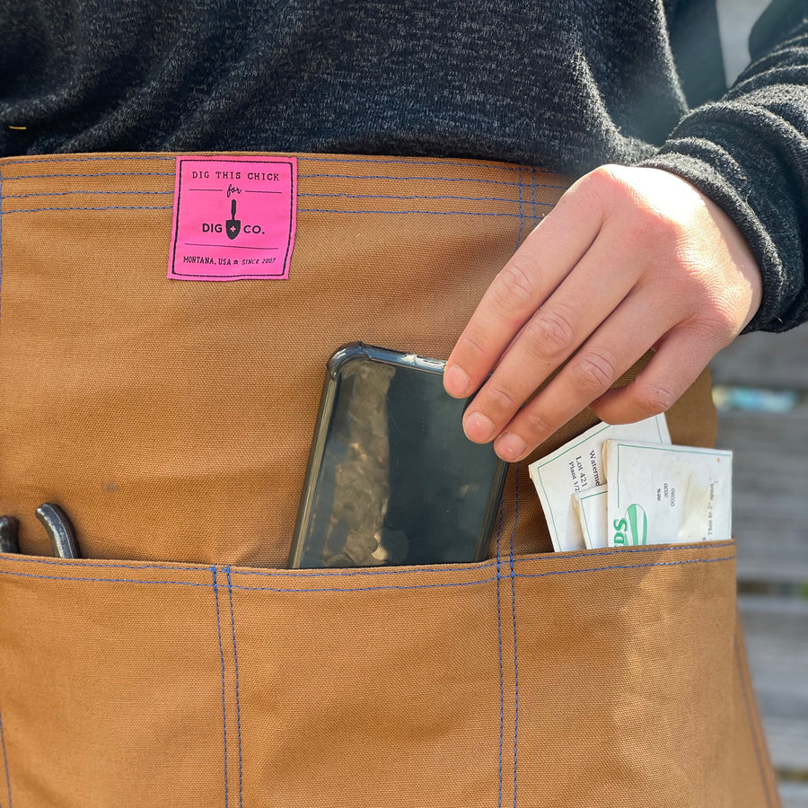 A person wears the canvas utility apron and pulls their phone out of the front pocket. 