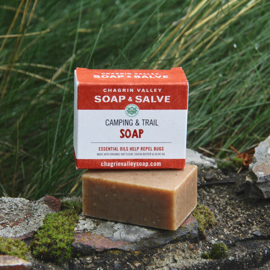A packaged bar of organic camp and trail soap that is also bug repellent. It is placed on a rock in front of grass. 