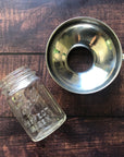 A stainless steel canning funnel and a glass jar are placed on a wooden surface. 