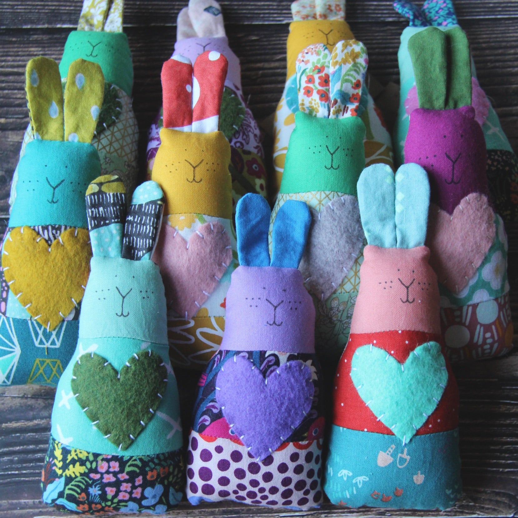 Eleven multicolored handmade fabric bunny rattles are lined up on wooden surface. 
