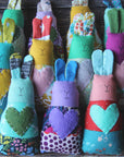 Eleven multicolored handmade fabric bunny rattles are lined up on wooden surface. 