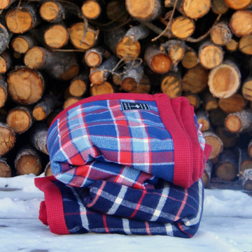Blue and red plaid flannel Camp Blanket folded and placed in the snow in front of a wood pile.