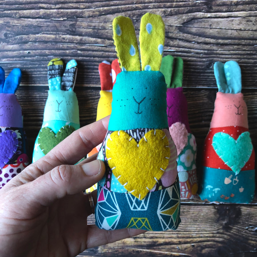 In the foreground, a hand holds up a blue and yellow handmade fabric bunny rattle, in the background there are five multicolored bunny rattles on a wooden surface.