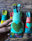 A hand holds a blue and green handmade bunny rattle in the foreground. In the background there are six multicolored bunny rattles placed in a line on a wooden background. 