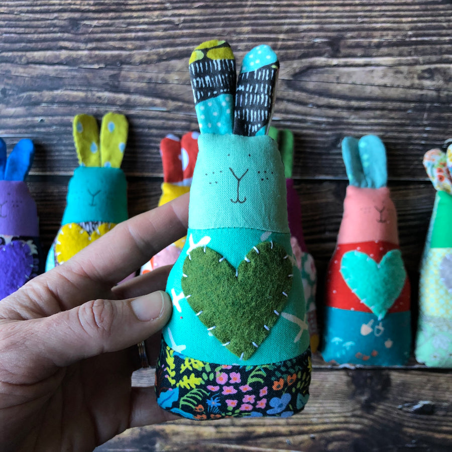 A hand holds a blue and green handmade bunny rattle in the foreground. In the background there are six multicolored bunny rattles placed in a line on a wooden background. 