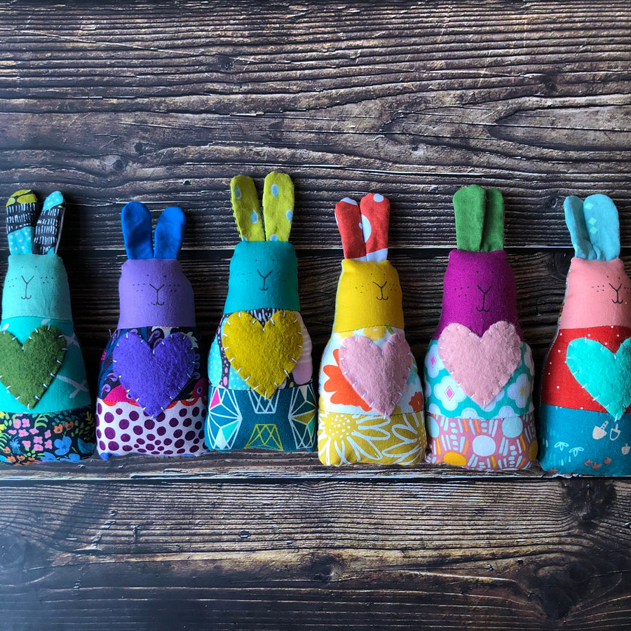 Six multicolored handmade fabric bunny rattles lie on a wooden surface. 