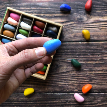 A hand holds a blue crayon rock. Crayon rocks are placed on a wooden surface. 