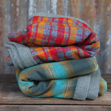 Two thick folded flannel camp blankets in blue plaid and red plaid are placed on a wooden board in front of a gray background. 