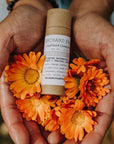 Brown compostable tube of pearl luster brightening balm, held in two hands accompanied by orange flowers 