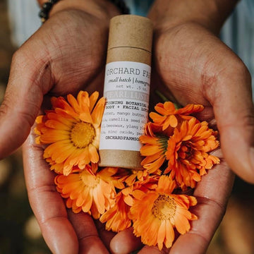 Brown compostable tube of pearl luster brightening balm, held in two hands accompanied by orange flowers 