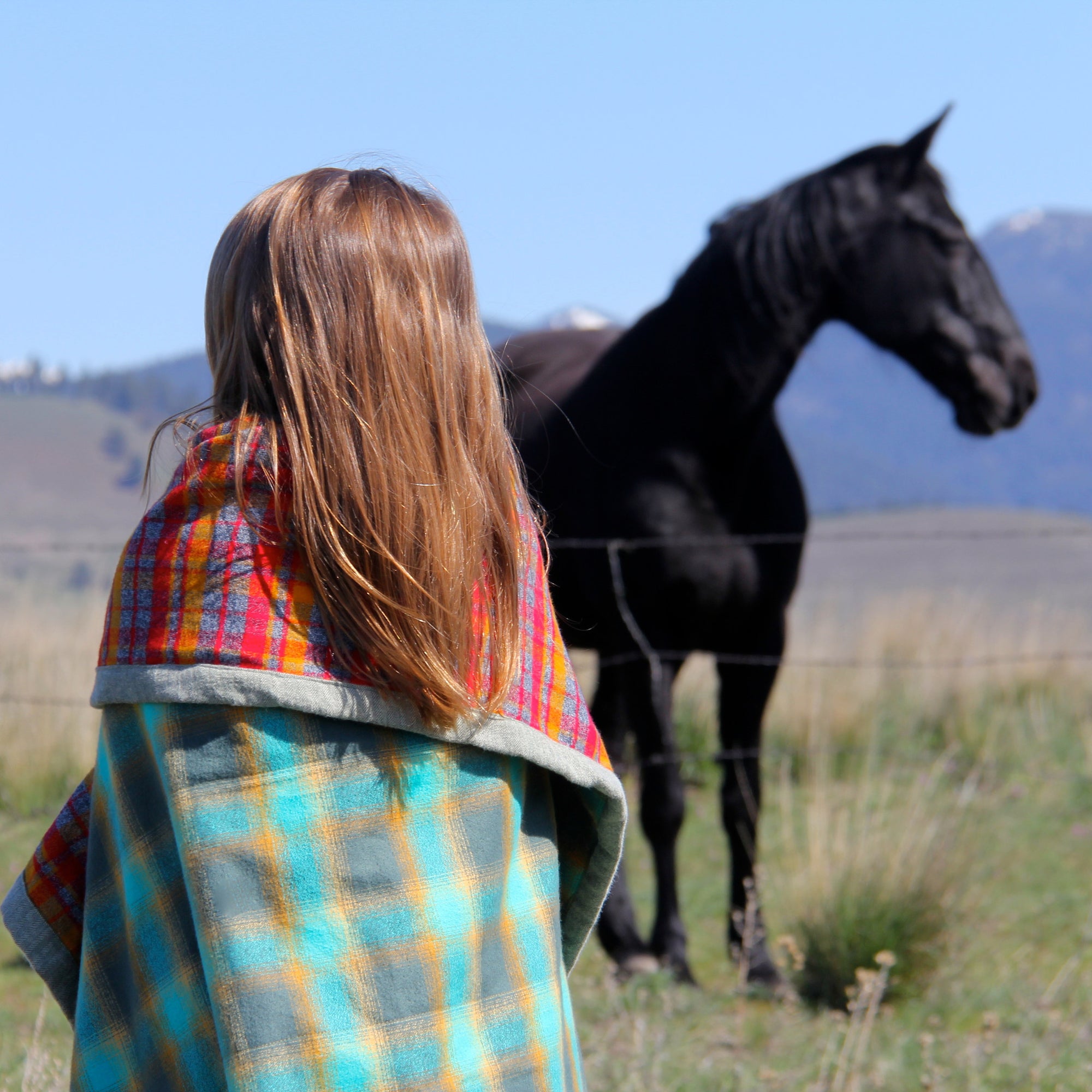 A young girl with loose brown hair stands with a Camp Blanket around her shoulders. A black horse stands in the background in a grassy field. 