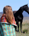 A young girl with loose brown hair stands with a Camp Blanket around her shoulders. A black horse stands in the background in a grassy field. 