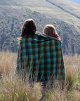 Two young girls wrapped in a blue and red plaid camp blanket stand in a grassy field in front of a grassy canyon and hill. 