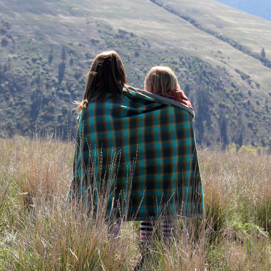 Two young girls wrapped in a blue and red plaid camp blanket stand in a grassy field in front of a grassy canyon and hill. 