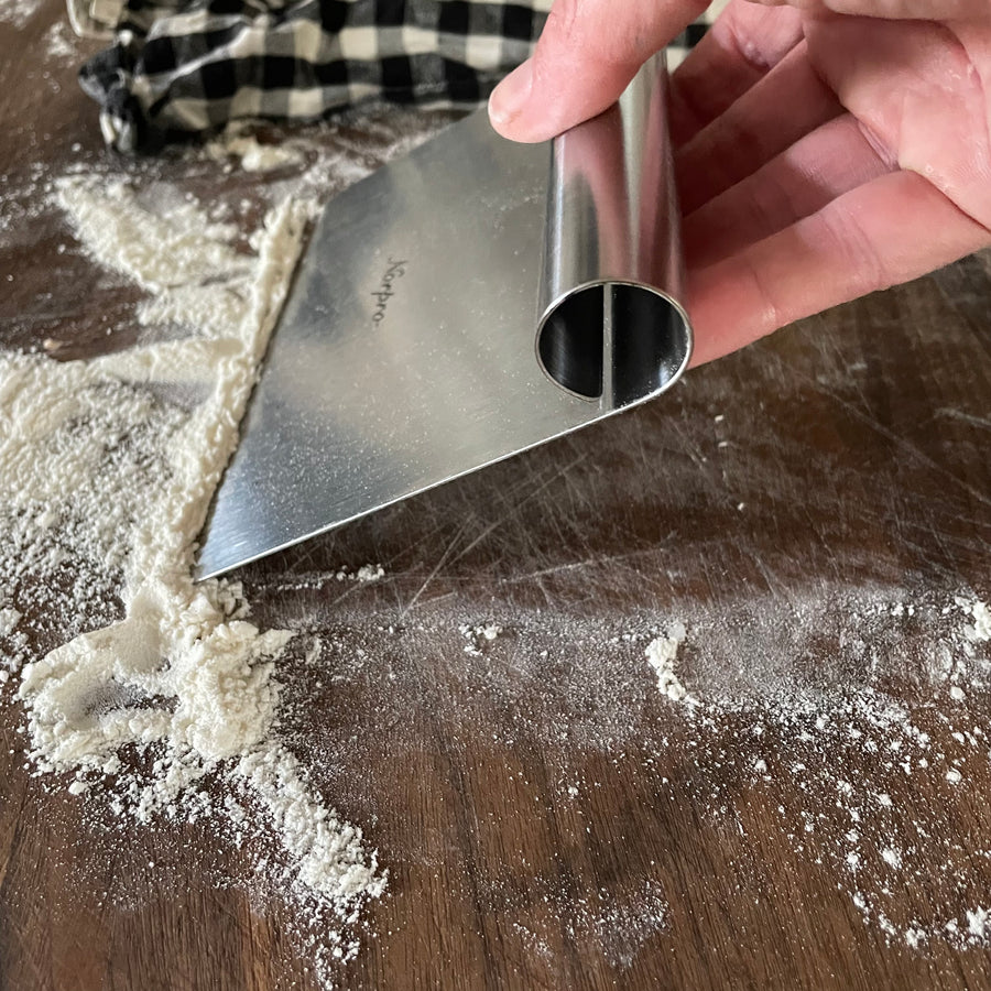 A hand demonstrates the use of a bench knife, scraping flour from a countertop
