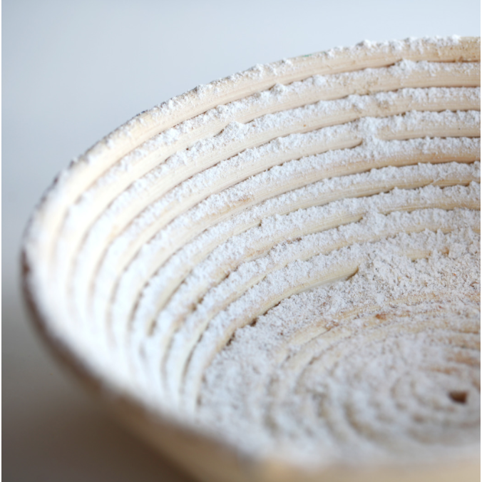 A close up of a floured bread proofing basket is displayed with a white background. 