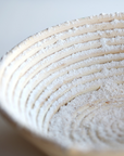 A close up of a floured bread proofing basket is displayed with a white background. 