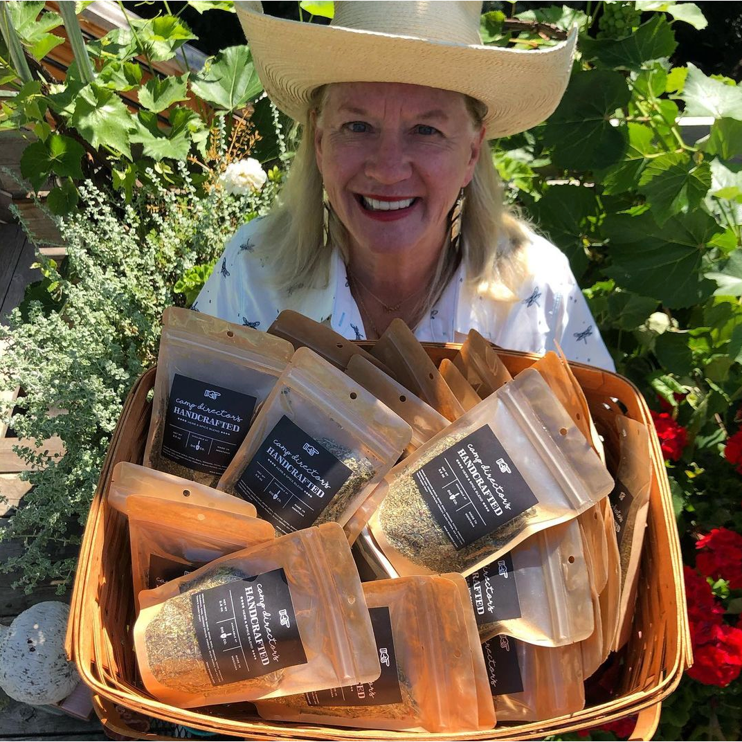 Terri Holt, standing in a garden,  looking up at the camera holding a basket of her handcrafted herb and spice blend.