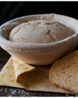 Risen bread dough sits in a bread proofing basket with a muslin liner on top of a yellow kitchen towel. Sliced bread sits next to the basket. 