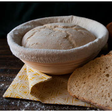 Risen bread dough sits in a bread proofing basket with a muslin liner on top of a yellow kitchen towel. Sliced bread sits next to the basket. 