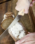 A hand cuts a loaf of rye bread with a serrated bread knife on a wooden cutting board. 