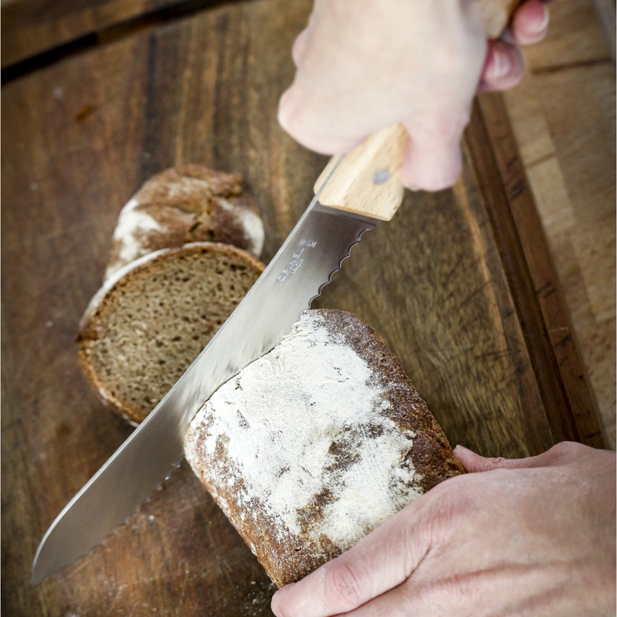 A hand cuts a loaf of rye bread with a serrated bread knife on a wooden cutting board. 