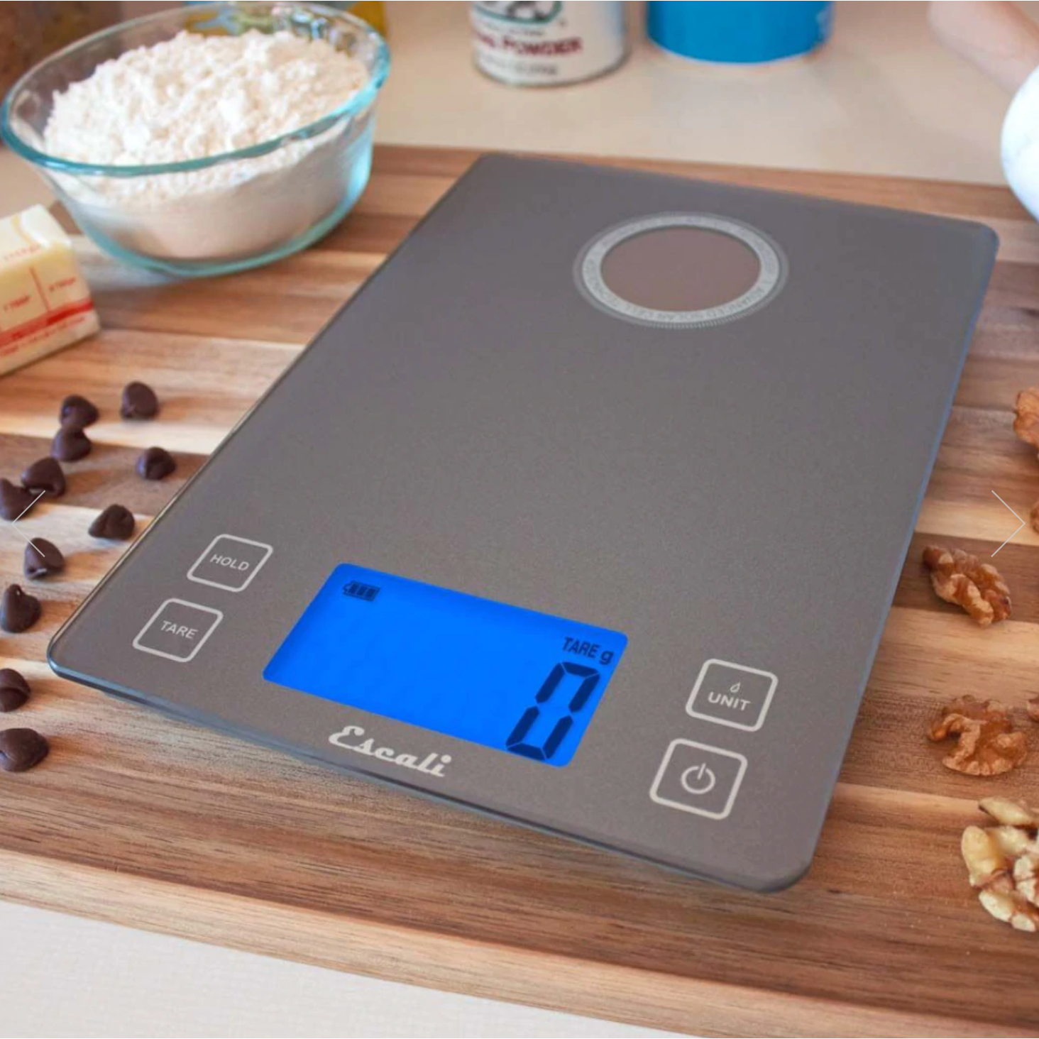 A silver-gray kitchen scale that is powered on sits on a wooden cutting board with pecans and chocolate chips scattered on top of it. A cup of flour, stick of butter, and a jar of baking powder are out of focus in the background. 