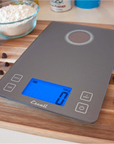 A silver-gray kitchen scale that is powered on sits on a wooden cutting board with pecans and chocolate chips scattered on top of it. A cup of flour, stick of butter, and a jar of baking powder are out of focus in the background. 