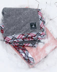 Two gray and pink handmade blankets with flannel trim are folded and lie in the snow. 