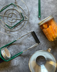 Contents of the Canning Gift Bundle displayed on a gray surface. Jar lifter, lid lifter, canning rack, jar with pickled carrots, and canning funnel. 