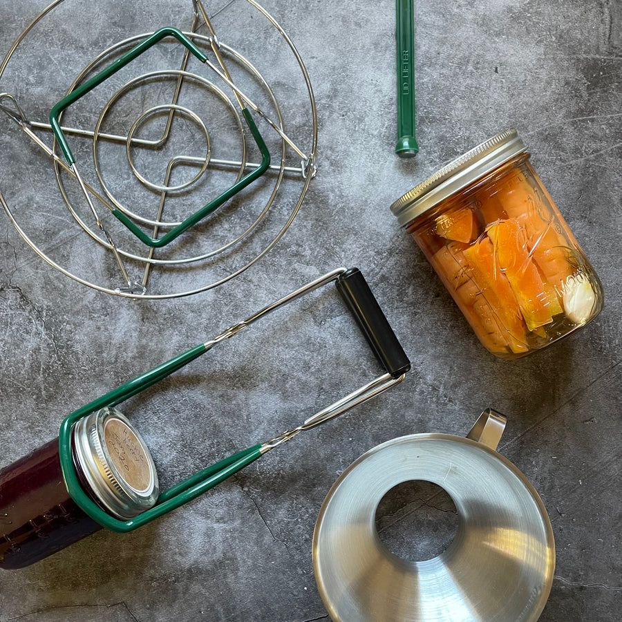 Contents of the Canning Gift Bundle displayed on a gray surface. Jar lifter, lid lifter, canning rack, jar with pickled carrots, and canning funnel. 