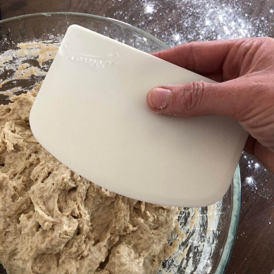 A hand holds a white bowl scraper over a glass bowl full of dough