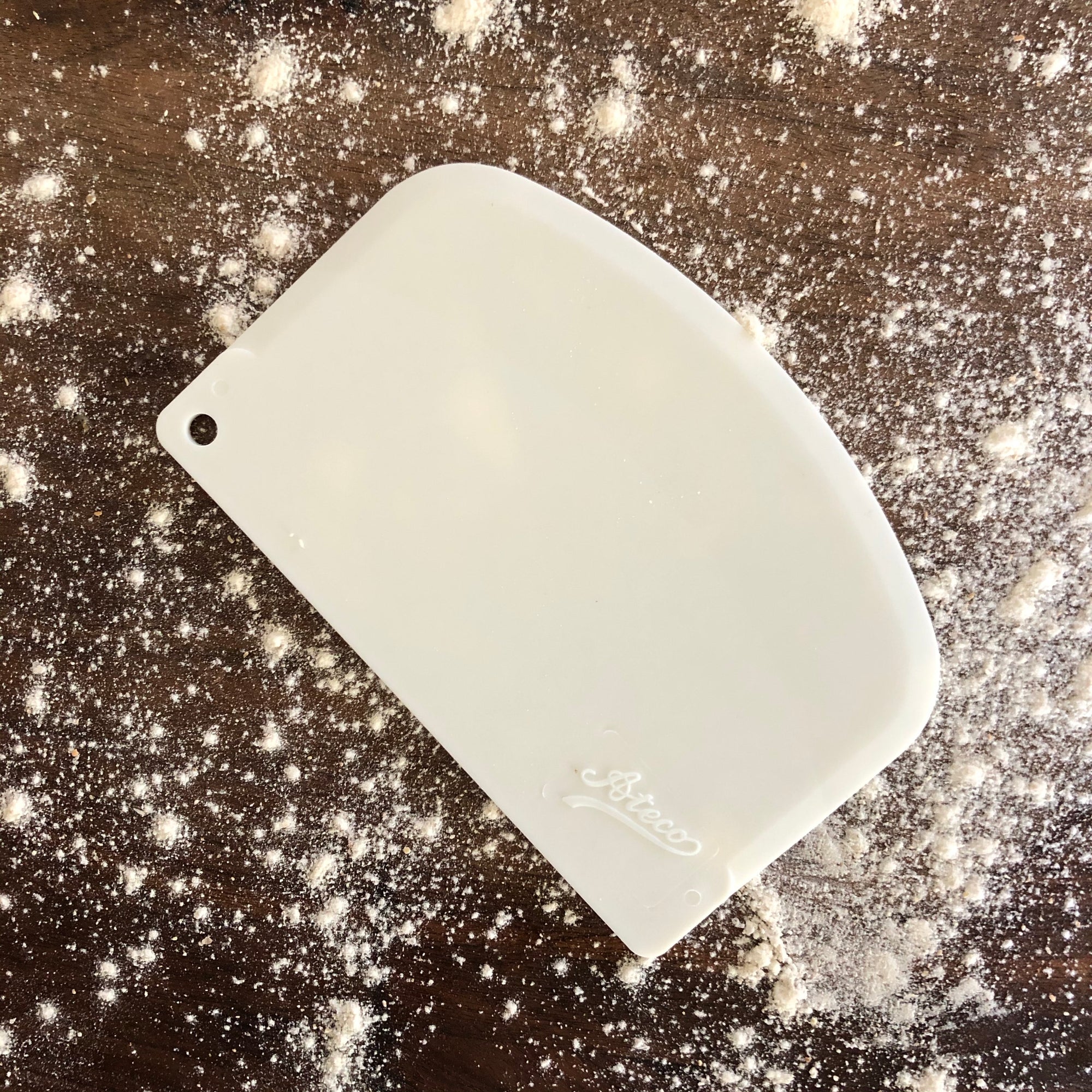 A white bowl scraper placed on a counter top covered in flour