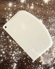 A white bowl scraper placed on a counter top covered in flour
