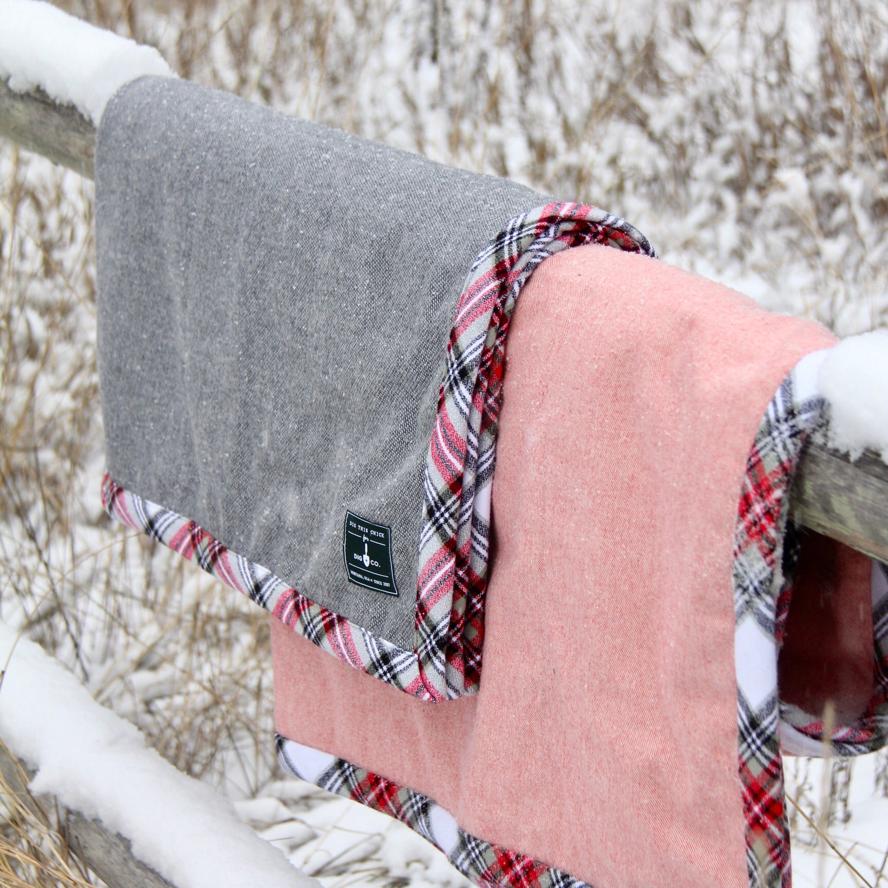 Two gray and pink blankets with flannel trim are hung over a wooden fence in the snow. 