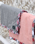 Two gray and pink blankets with flannel trim are hung over a wooden fence in the snow. 