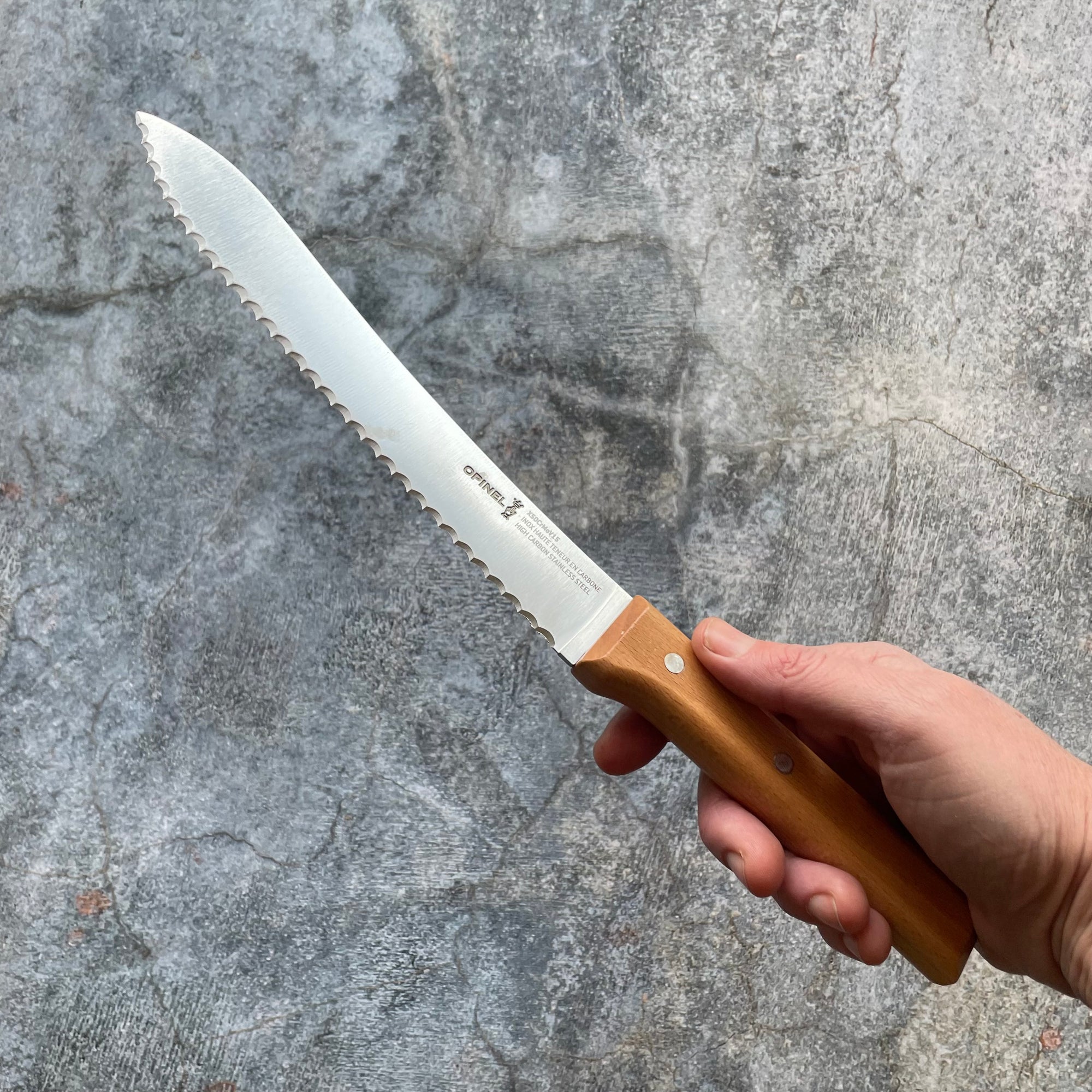 A hand holds a serrated bread knife with a wooden handle in front of a gray backdrop. 