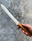A hand holds a serrated bread knife with a wooden handle in front of a gray backdrop. 