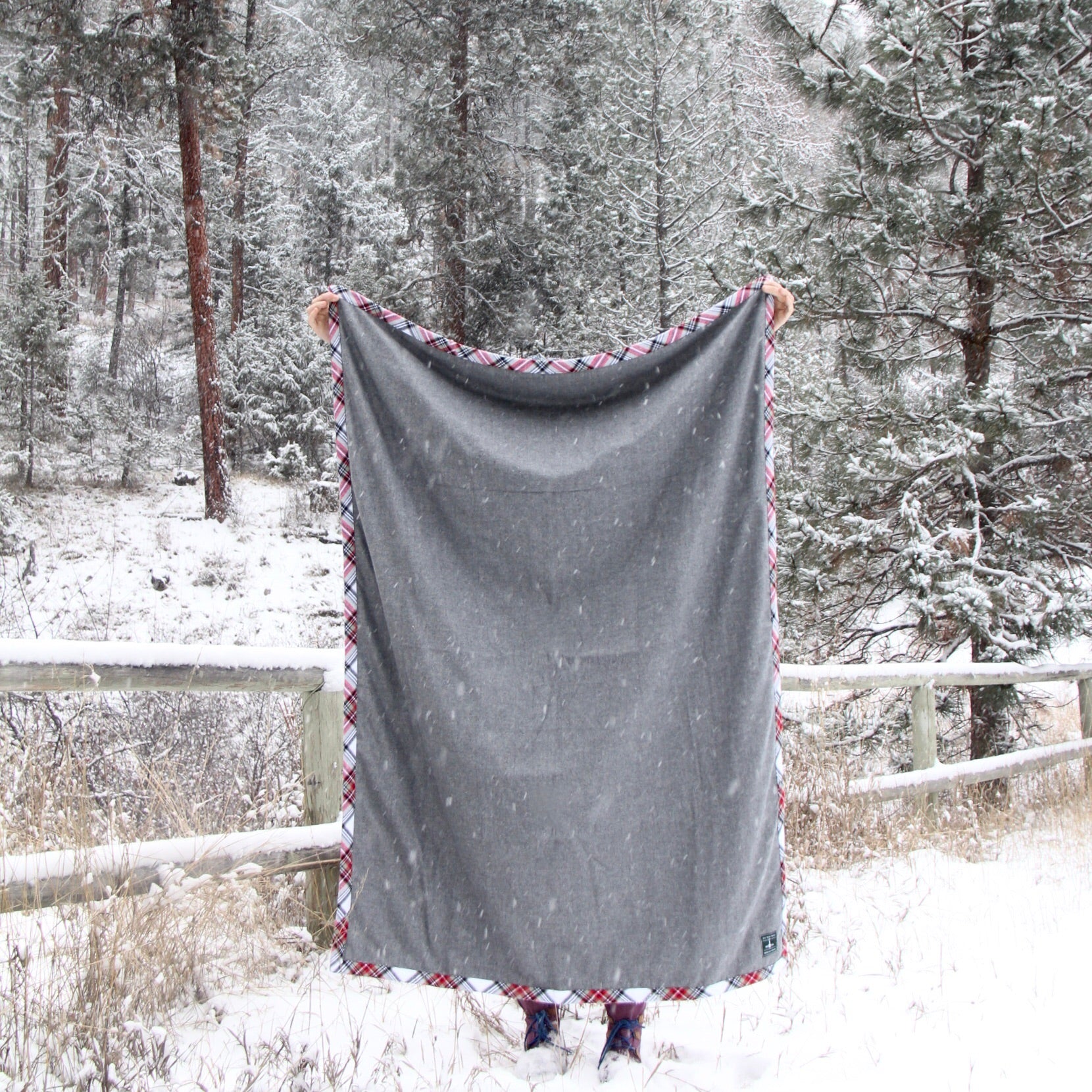 A person holds a gray blanket with flannel trim in front of them in the snow. Snow covered trees and a wooden fence are in the background. 